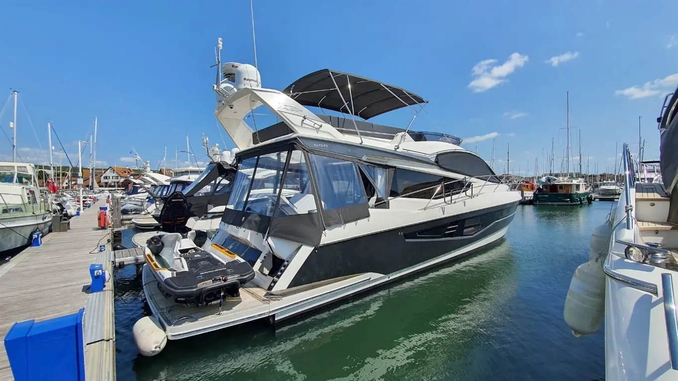 Luxury yacht docked at marina, featuring sleek design, large windows, and a jet ski on board, with boats and blue sky in background.